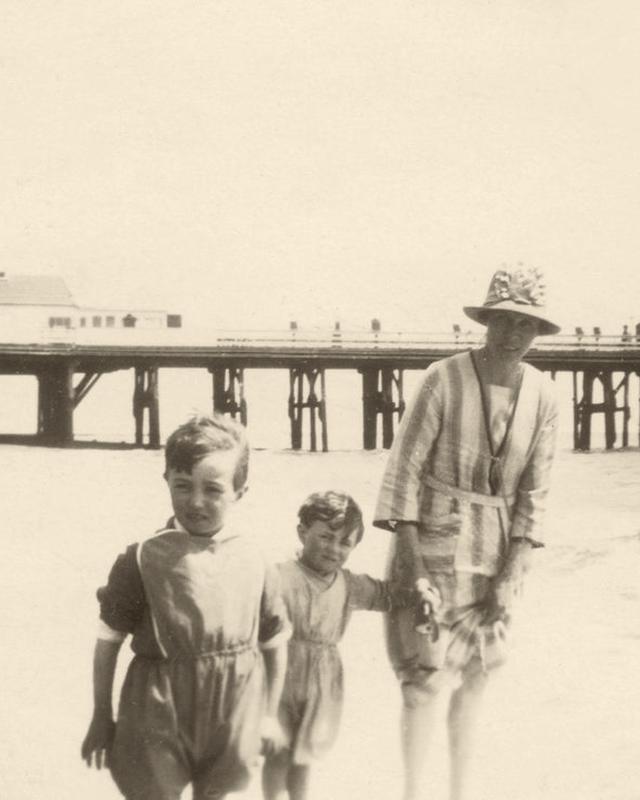 A sepia photograph of a family on a beach beside a pier in the early nineteen sixties
